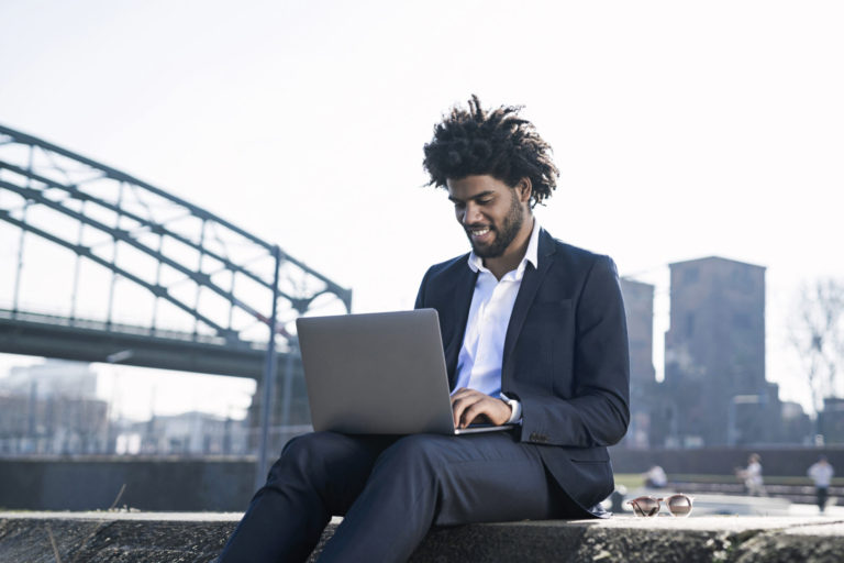 Smiling businessman sitting at the riverside using laptop