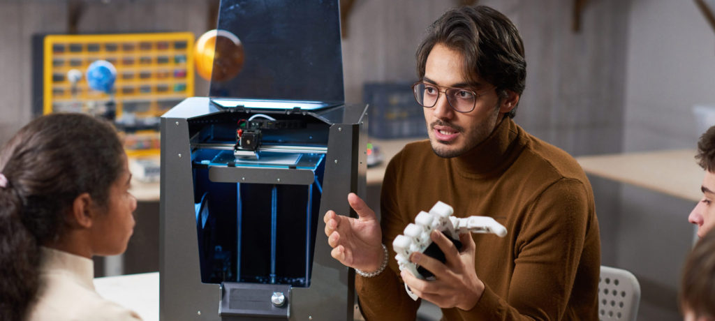 Young man in eyeglasses holding robotic arm and talking to his students about the artificial intelligence during science lesson